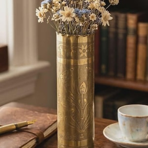 May include: A brass vase with engraved floral designs, holding dried lavender and white and blue flowers. The vase is on a wooden table with a notebook, pen, and teacup. The text on the vase reads: 1916 TRENSCH ART SHELL CASING, ENGRAVED BY HAND, UNIT: REMBACH.