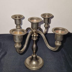 May include: A silver-colored candelabra with five candle holders. The candelabra has a decorative base and curved arms. The candle holders are circular and have a slightly reddish interior. The candelabra is made of metal.