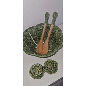Peut inclure: Un saladier vert en céramique en forme de chou avec deux couverts à salade en bois. Le bol a une surface texturée et un motif de veines blanches. Un plat assorti à deux sections est en dessous.