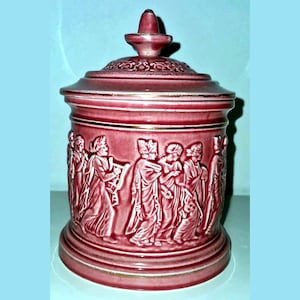 Op de afbeelding: Een cilindrische keramische pot met bijpassend deksel, voorzien van een dieproze glazuur en goudkleurige randen. De buitenkant is versierd met een doorlopend reliëf dat een processie van figuren in vloeiende gewaden toont, sommigen met instrumenten.