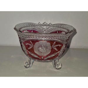 Peut inclure: Un bol en verre transparent et rouge canneberge présentant un motif en cristal taillé avec une rose gravée au centre. Le bol possède un bord festonné et repose sur trois petits pieds en verre transparent, affichant un design décoratif détaillé.