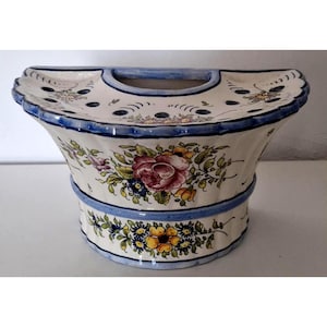 Op de afbeelding: Een keramische bloemenvaas met een blauwe rand en handvat. De vaas heeft een crèmekleurige basis met handgeschilderde bloemen in tinten rood, geel en blauw. De vaas heeft een unieke vorm met een brede bovenkant en een smallere basis.