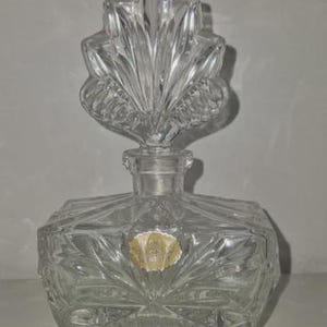 May include: A clear, vintage glass perfume bottle with a decorative leaf-shaped stopper. The bottle has a square base with a starburst design and is supported by small feet. A small, faded label is affixed to the front.
