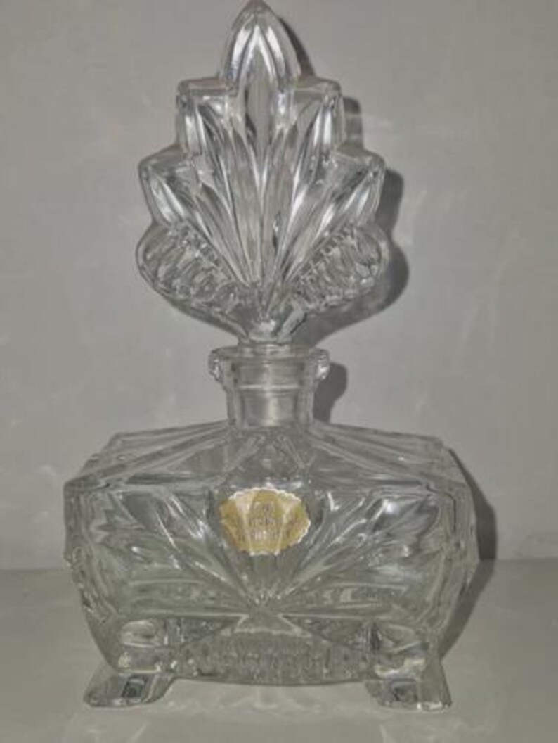 May include: A clear, vintage glass perfume bottle with a decorative leaf-shaped stopper. The bottle has a square base with a starburst design and is supported by small feet. A small, faded label is affixed to the front.