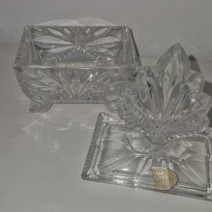 May include: A clear, cut-glass trinket box with a lid. The box is rectangular with a floral design and sits on small feet. The lid is shaped like a leaf. A small gold sticker is visible on the base.
