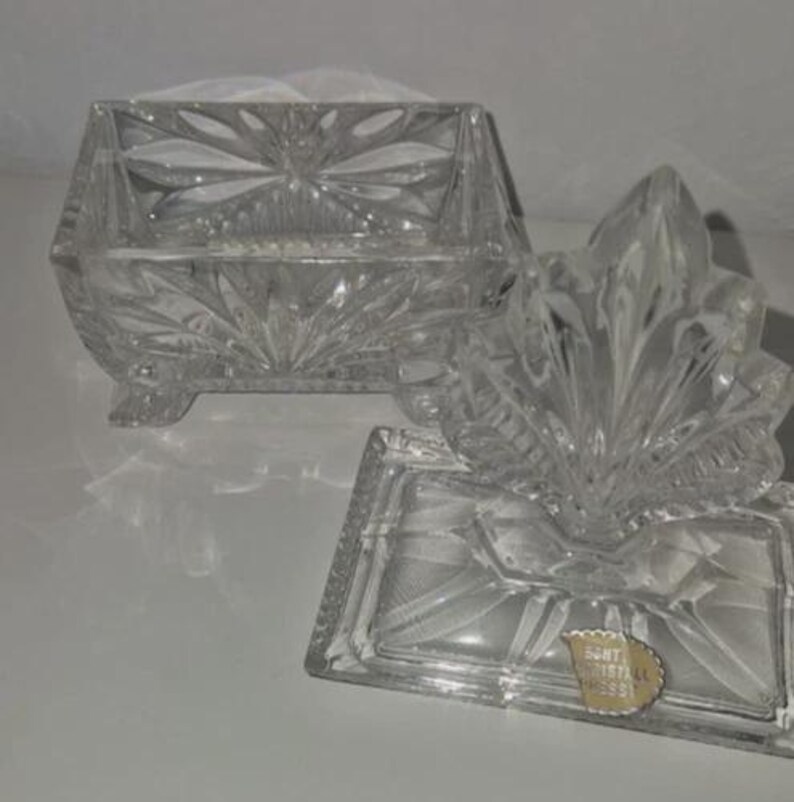 May include: A clear, cut-glass trinket box with a lid. The box is rectangular with a floral design and sits on small feet. The lid is shaped like a leaf. A small gold sticker is visible on the base.