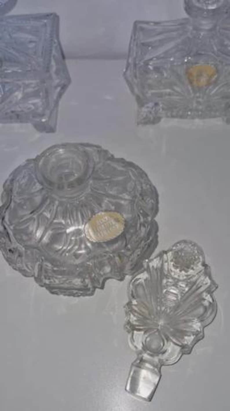 May include: Clear glass perfume bottles with intricate designs. One bottle is round with a floral pattern, and another has a rectangular shape. A decorative stopper is next to the round bottle. The bottles are empty and appear to be vintage.