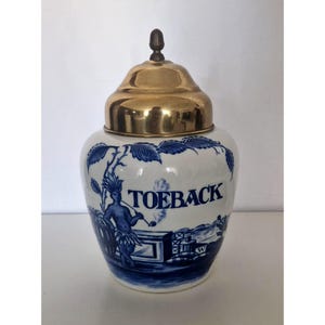 Pode incluir: Um pote de cerâmica branca com um padrão de transferência azul apresentando uma figura a fumar um cachimbo e o texto TOEBACK. O pote tem um corpo arredondado e uma tampa metálica removível em tom dourado com um pequeno remate no topo.