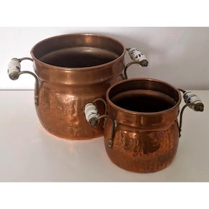 Op de afbeelding: Twee koperen potten met een gehamerde textuur. Elke pot heeft een afgeronde vorm en een brede opening. De potten hebben gebogen handvatten met witte keramische grepen versierd met blauwe bloempatronen. De potten zijn verschillend van grootte.