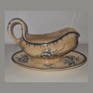 May include: Cream-colored gravy boat with a matching saucer. The boat has a curved handle and a spout. Both pieces are decorated with blue floral patterns and a blue trim along the edges. The set is made of ceramic.
