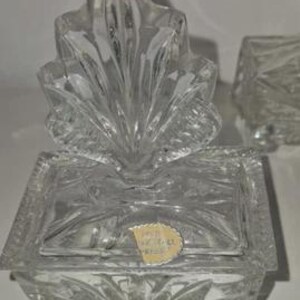 May include: A clear, cut-glass trinket box with a decorative lid. The box has a rectangular base with a lid and is adorned with a leaf-like design. A ruler is placed next to the box. The box is approximately 4 inches long.