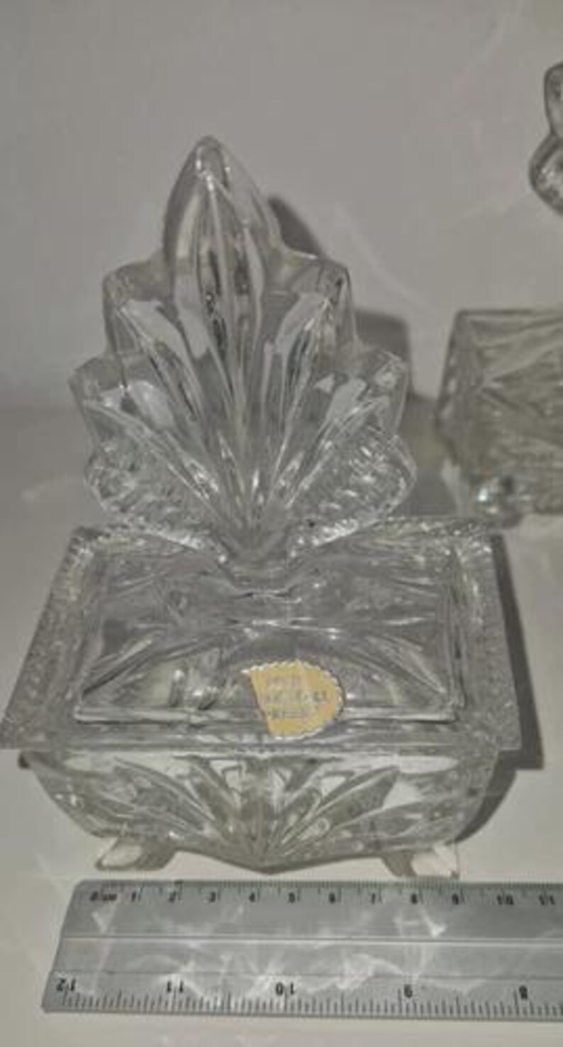 May include: A clear, cut-glass trinket box with a decorative lid. The box has a rectangular base with a lid and is adorned with a leaf-like design. A ruler is placed next to the box. The box is approximately 4 inches long.