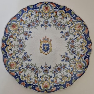 May include: A decorative ceramic plate with a scalloped edge. The plate features a white center with a floral and scroll design in blue, yellow, and peach. A crest with a crown and fleur-de-lis is in the center.