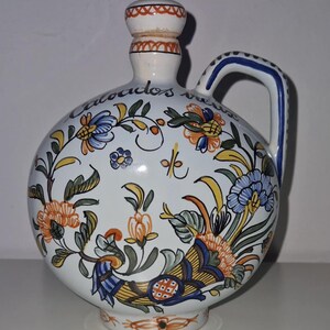 May include: A white ceramic pitcher with a round body and a handle. The pitcher is decorated with colorful floral patterns in blue, orange, and yellow. The neck and handle have blue accents. The text "calocados" is written on the pitcher.
