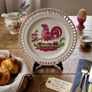 Pode incluir: Um prato de cerâmica vintage com um desenho de galo. O prato apresenta um fundo branco, uma borda ondulada bordô e uma ilustração de galo em tons de rosa e vermelho. O prato é exibido em um suporte preto.