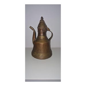 May include: A weathered copper-toned metal pitcher featuring a wide, tapered base, a slender curved spout, and a pointed lid. The vessel has a prominent handle attached to the side and displays a dull, aged patina across its entire exterior surface.