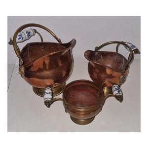 Peut inclure: Un ensemble de trois récipients en cuivre dotés de poignées en laiton avec des accents en céramique blanche et bleue. La collection comprend deux grands seaux à anse mobile et un bol plus petit à deux anses latérales, sur socles en laiton.