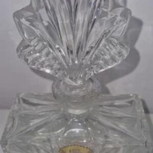 May include: A clear, vintage perfume bottle with a decorative, leaf-shaped stopper. The bottle has a rectangular base and is made of cut glass. A gold-colored label is affixed to the front of the bottle. The bottle is designed to sit on a flat surface.