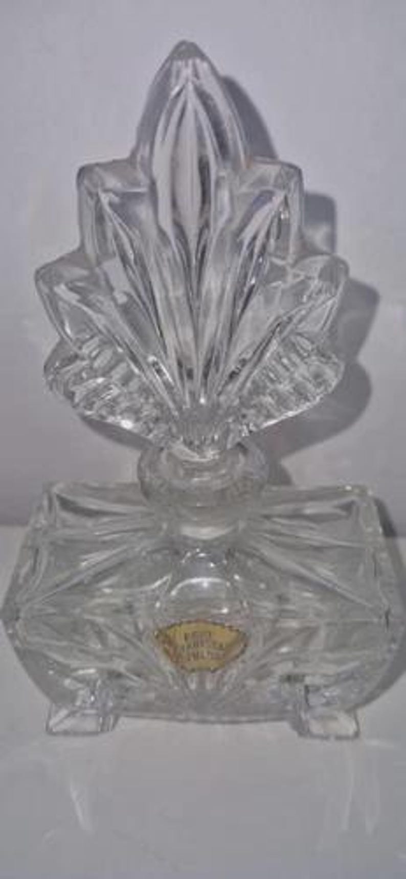 May include: A clear, vintage perfume bottle with a decorative, leaf-shaped stopper. The bottle has a rectangular base and is made of cut glass. A gold-colored label is affixed to the front of the bottle. The bottle is designed to sit on a flat surface.