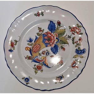 May include: White ceramic plate with a scalloped edge and a blue rim. The plate features a central floral design with red, blue, yellow, and green colours. Additional floral motifs are scattered around the plate's edge.