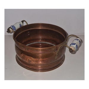 Può includere: Una bacinella rotonda in rame dotata di due manici in ottone con impugnature in ceramica bianca. Ogni impugnatura in ceramica è decorata con un motivo floreale blu. Il recipiente in rame ha una finitura liscia e lucida e un'apertura ampia.