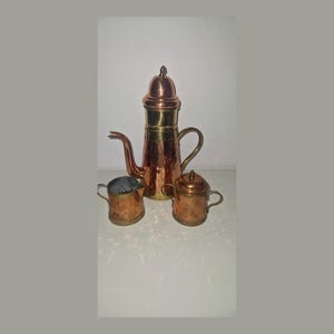 Peut inclure: Une grande cafetière en cuivre et laiton avec un bec verseur et une anse incurvés, accompagnée d'un pot à lait et d'un sucrier assortis avec couvercle. Chaque pièce présente une texture martelée et une finition polie, soulignant le contraste.