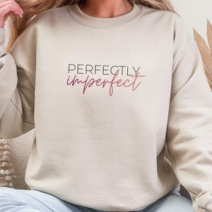 Puede incluir: Sudadera beige de cuello redondo con el texto "Perfectly Imperfect" en letras negras y rosas. La sudadera es de color neutro y tiene mangas largas. La modelo lleva vaqueros azul claro.