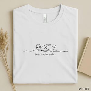 Könnte beinhalten: Weißes T-Shirt mit Rundhalsausschnitt und einer minimalistischen Strichzeichnung eines Schwimmers mit Brille und Badekappe. Der Text "Water is my happy place" befindet sich unter der Zeichnung.