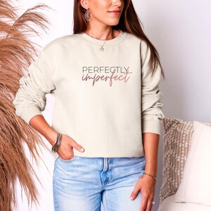 May include: Cream-colored sweatshirt with the words "Perfectly Imperfect" in a script font. The long-sleeved top is a neutral color and is paired with blue jeans. The sweatshirt is a casual style.