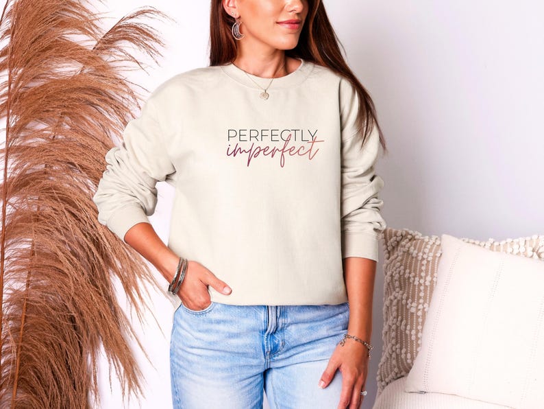 May include: Cream-colored sweatshirt with the words "Perfectly Imperfect" in a script font. The long-sleeved top is a neutral color and is paired with blue jeans. The sweatshirt is a casual style.