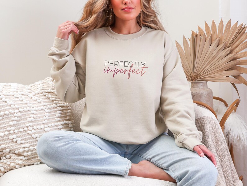 May include: Beige crewneck sweatshirt with the text "Perfectly Imperfect" in black and pink script. The sweatshirt is a neutral color and has long sleeves. The model is wearing light blue jeans.