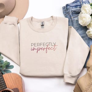 May include: A beige sweatshirt with the text "Perfectly imperfect" in a burgundy script. The sweatshirt is laid out with a guitar, a hat, jeans, boots, and flowers.