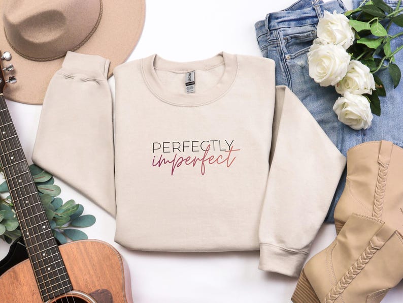 May include: A beige sweatshirt with the text "Perfectly imperfect" in a burgundy script. The sweatshirt is laid out with a guitar, a hat, jeans, boots, and flowers.