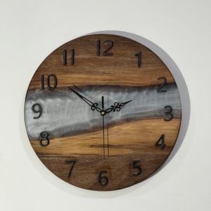 Könnte beinhalten: Runde Wanduhr aus Holz mit natürlicher Holzmaserung und einem zentralen Band aus schimmerndem, perlmuttartigem Harz. Schwarze Ziffern markieren die Stunden, und die Zeiger der Uhr sind schwarz. Die Uhr ist an einer weißen Wand befestigt.