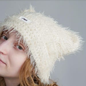 May include: A cream-colored, fuzzy, textured beanie hat with a pointed cat-ear shape. The hat features a small white rectangular fabric label on the front with the black text ZAIKA printed on it. The material has a long, soft, shaggy pile.