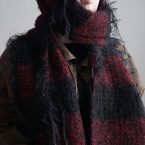 May include: A fuzzy, textured scarf featuring a bold, alternating plaid pattern in deep black and dark red. The scarf has a loose, shaggy knit construction with visible frayed edges and is wrapped around the head and neck over a brown jacket.