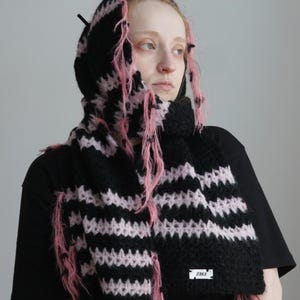 May include: A black and light pink striped knit hooded scarf featuring a fuzzy, textured yarn construction with loose, frayed pink threads throughout. A small white rectangular label with the black text ZAKA is attached to the lower edge.