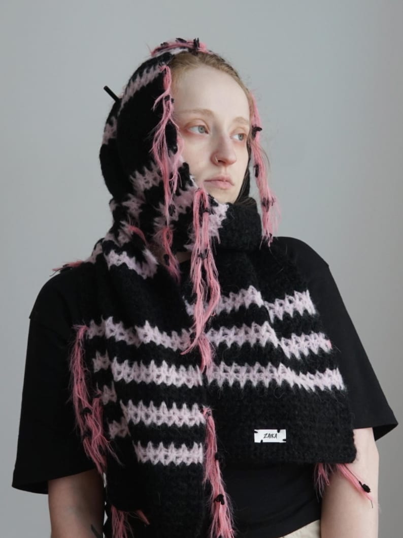 May include: A black and light pink striped knit hooded scarf featuring a fuzzy, textured yarn construction with loose, frayed pink threads throughout. A small white rectangular label with the black text ZAKA is attached to the lower edge.