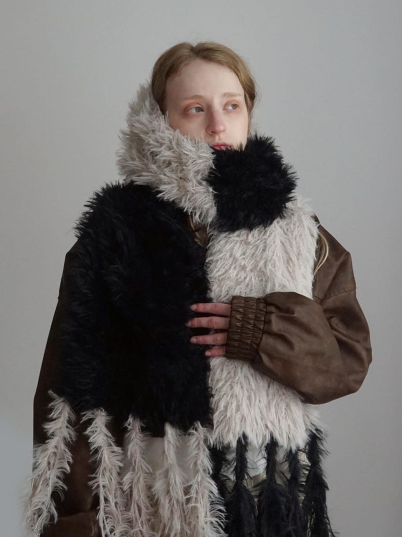 May include: A long, textured scarf featuring alternating vertical panels of black and light gray faux fur. The scarf is designed with long, shaggy fringe detailing at the ends, incorporating both black and light gray strands throughout the design.