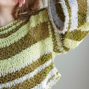 May include: A long-sleeved crochet jumper featuring a horizontal striped pattern in shades of olive green, light lime green, and soft lavender. The garment is constructed with an open, airy knit texture and wide sleeves, showcasing a loose, semi-sheer weave.