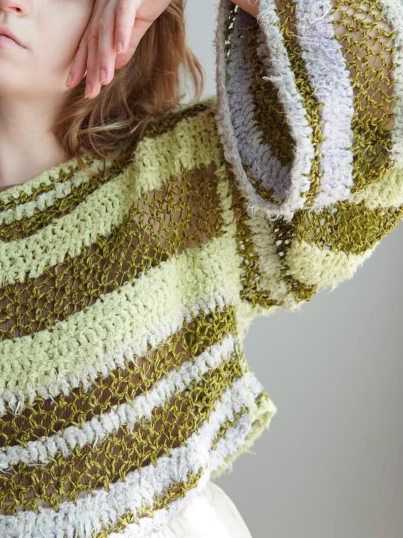 May include: A long-sleeved crochet jumper featuring a horizontal striped pattern in shades of olive green, light lime green, and soft lavender. The garment is constructed with an open, airy knit texture and wide sleeves, showcasing a loose, semi-sheer weave.