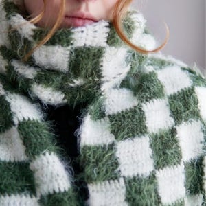 May include: A crochet scarf featuring a checkered pattern in alternating dark forest green and off-white squares. The yarn has a fuzzy, textured appearance, and the scarf is wrapped around the neck with a high, thick collar construction.