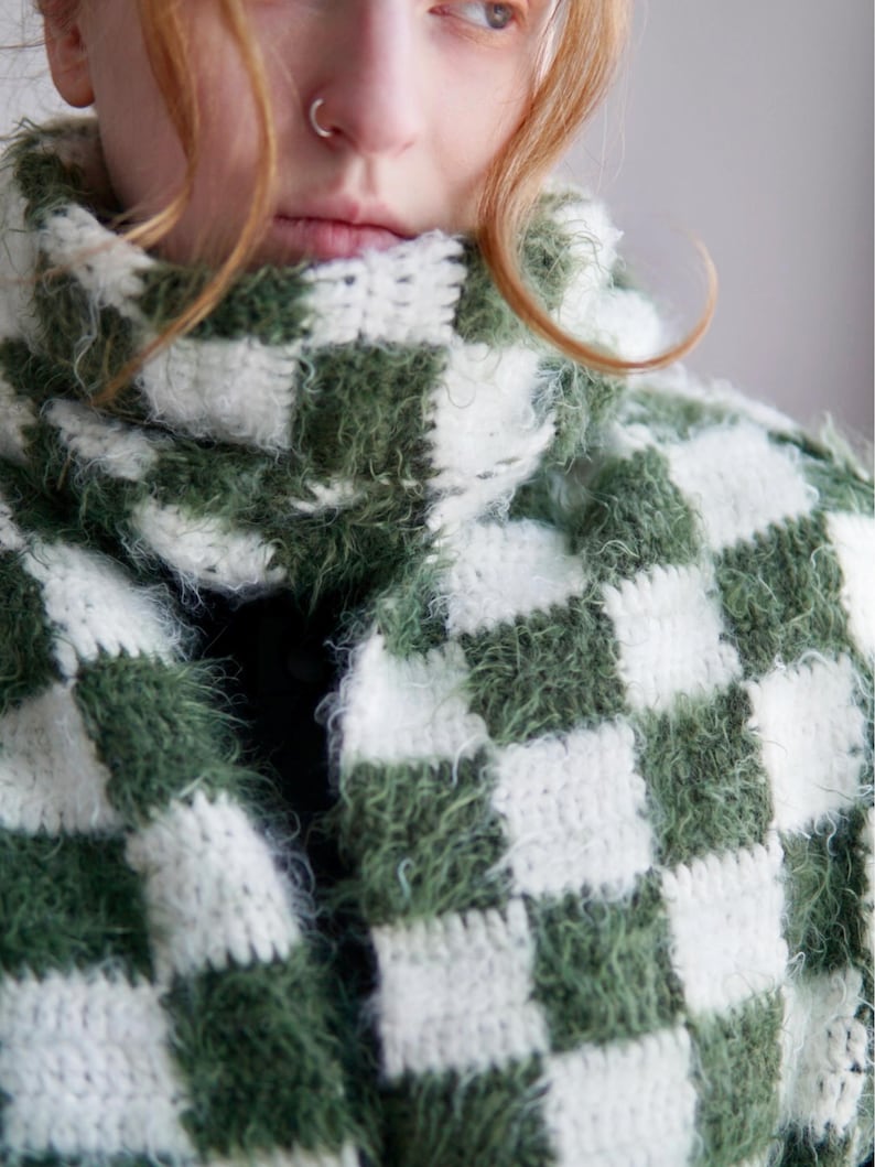 May include: A crochet scarf featuring a checkered pattern in alternating dark forest green and off-white squares. The yarn has a fuzzy, textured appearance, and the scarf is wrapped around the neck with a high, thick collar construction.
