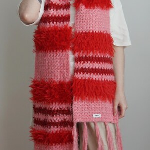 May include: A long, rectangular crochet scarf featuring horizontal stripes in shades of red, dark red, and light pink. The scarf incorporates alternating sections of smooth yarn and textured, shaggy yarn, finished with long pink fringe at the ends.