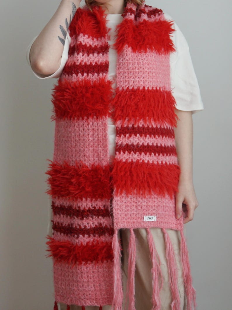 May include: A long, rectangular crochet scarf featuring horizontal stripes in shades of red, dark red, and light pink. The scarf incorporates alternating sections of smooth yarn and textured, shaggy yarn, finished with long pink fringe at the ends.