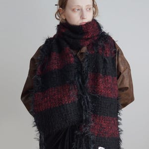 May include: A long, textured scarf featuring wide horizontal stripes in alternating black and deep red. The knit fabric has a fuzzy, shaggy appearance with loose fibres along the edges. A small white rectangular label is attached to the bottom corner.