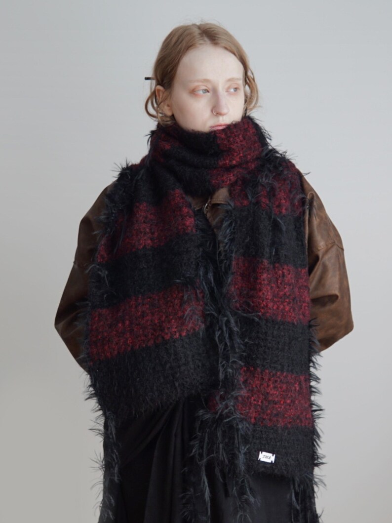 May include: A long, textured scarf featuring wide horizontal stripes in alternating black and deep red. The knit fabric has a fuzzy, shaggy appearance with loose fibres along the edges. A small white rectangular label is attached to the bottom corner.