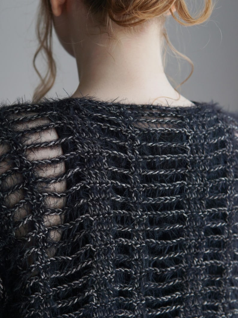 May include: A dark charcoal gray knit top featuring a loose, open-weave construction with a fuzzy, textured yarn. The garment displays a repeating vertical pattern of horizontal stitches, creating a semi-sheer effect across the back and shoulders.