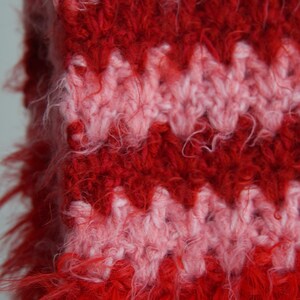 May include: A crochet fabric featuring a repeating chevron pattern in alternating horizontal stripes of vibrant red and soft pink. The yarn has a fuzzy, textured appearance with loose fibres extending from the surface of the knitted stitches.