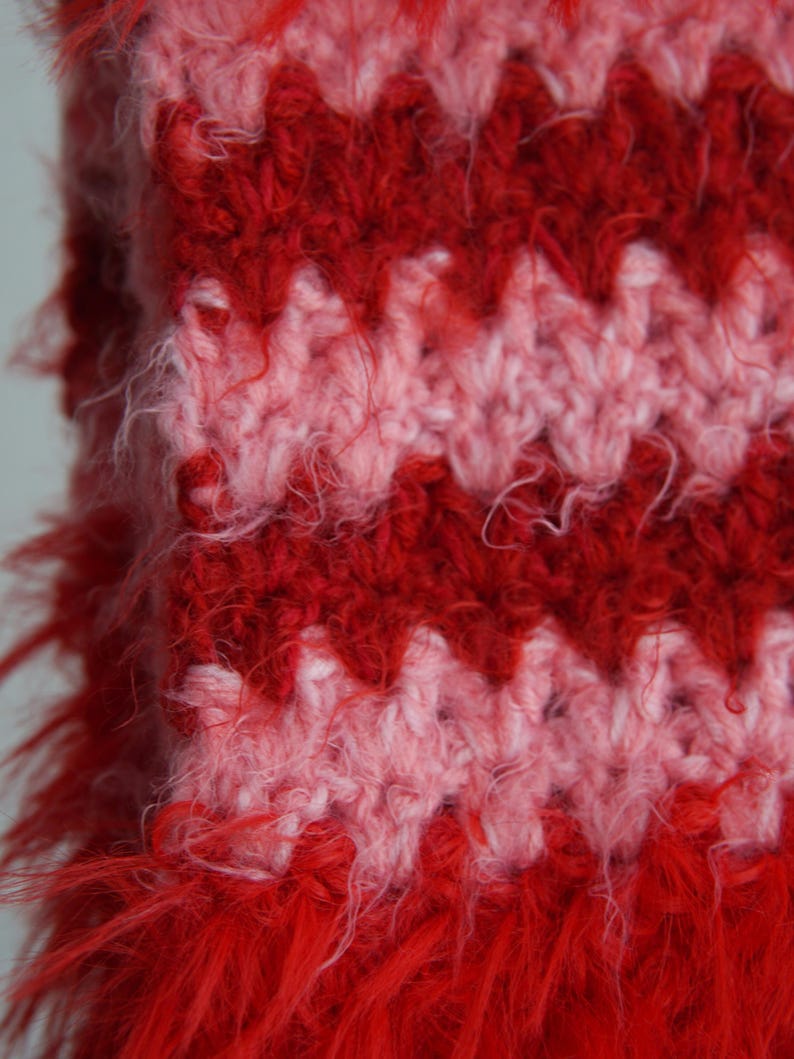 May include: A crochet fabric featuring a repeating chevron pattern in alternating horizontal stripes of vibrant red and soft pink. The yarn has a fuzzy, textured appearance with loose fibres extending from the surface of the knitted stitches.